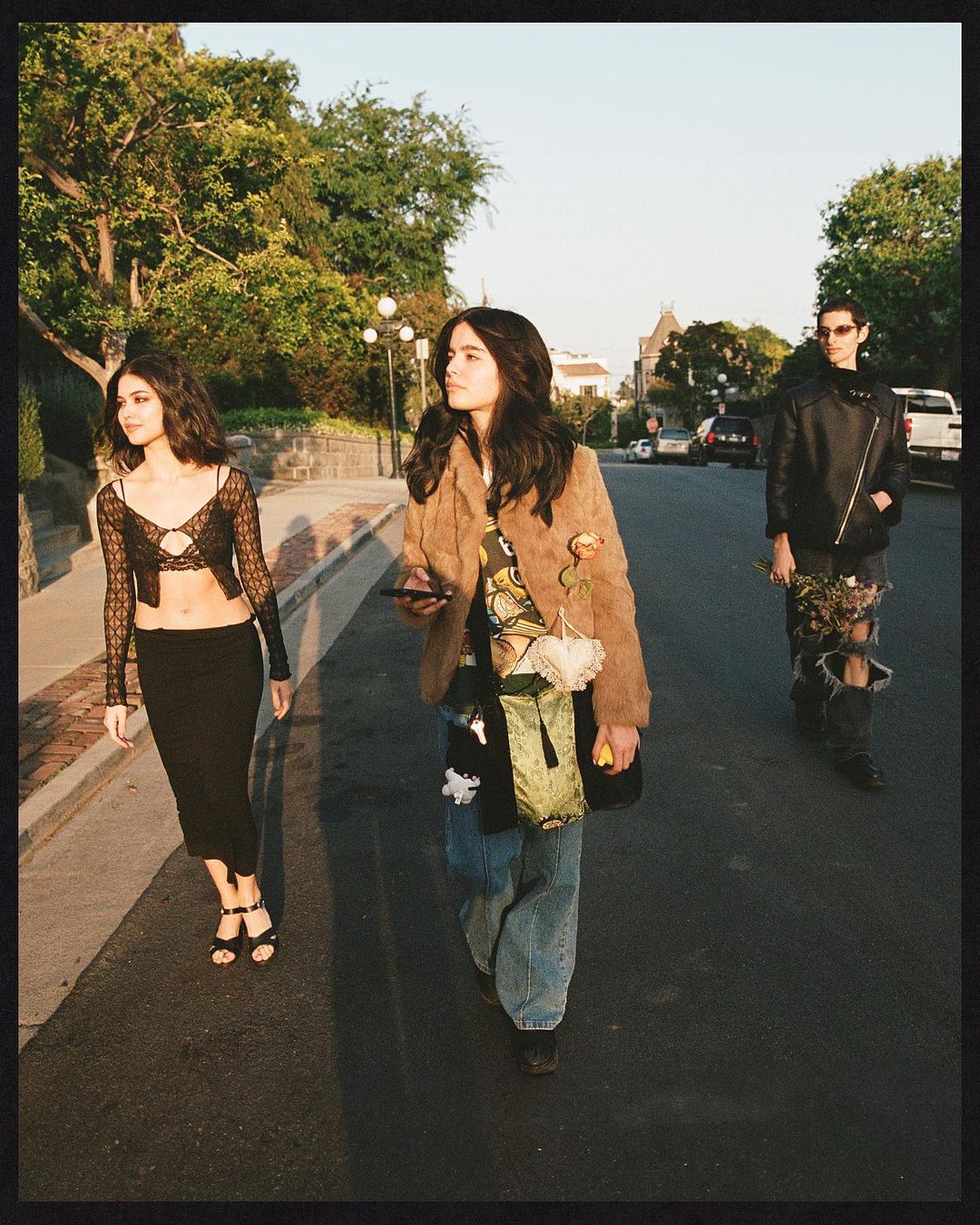 Three stylish young adults walk down a tree-lined street on a sunny day; the woman in the middle looks at her phone.