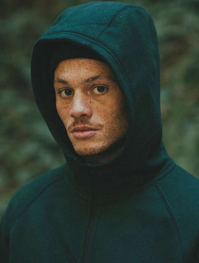 Close-up portrait of an adult male wearing a dark green hooded sweatshirt, eyes forward, with a blurred green natural background.