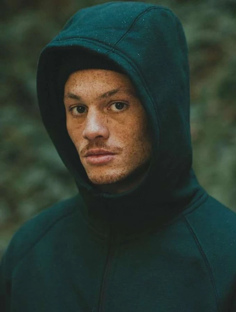 Close-up portrait of an adult male wearing a dark green hooded sweatshirt, eyes forward, with a blurred green natural background.
