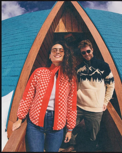 Couple posing in front of A-frame building with blue roof. Woman wears red checkered jacket and sunglasses; man wears mountain sweater.