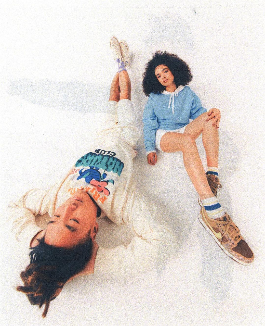 Two young adults in casual streetwear pose against a white backdrop. The man lies on his back while the woman sits with crossed legs.