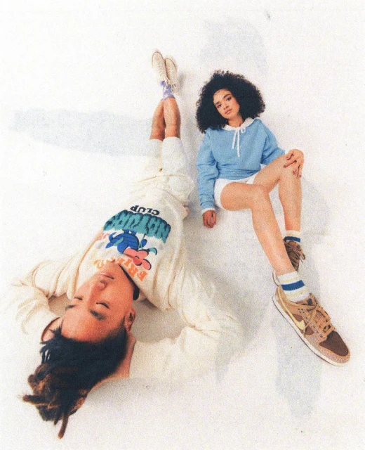 Two young adults in casual streetwear pose against a white backdrop. The man lies on his back while the woman sits with crossed legs.