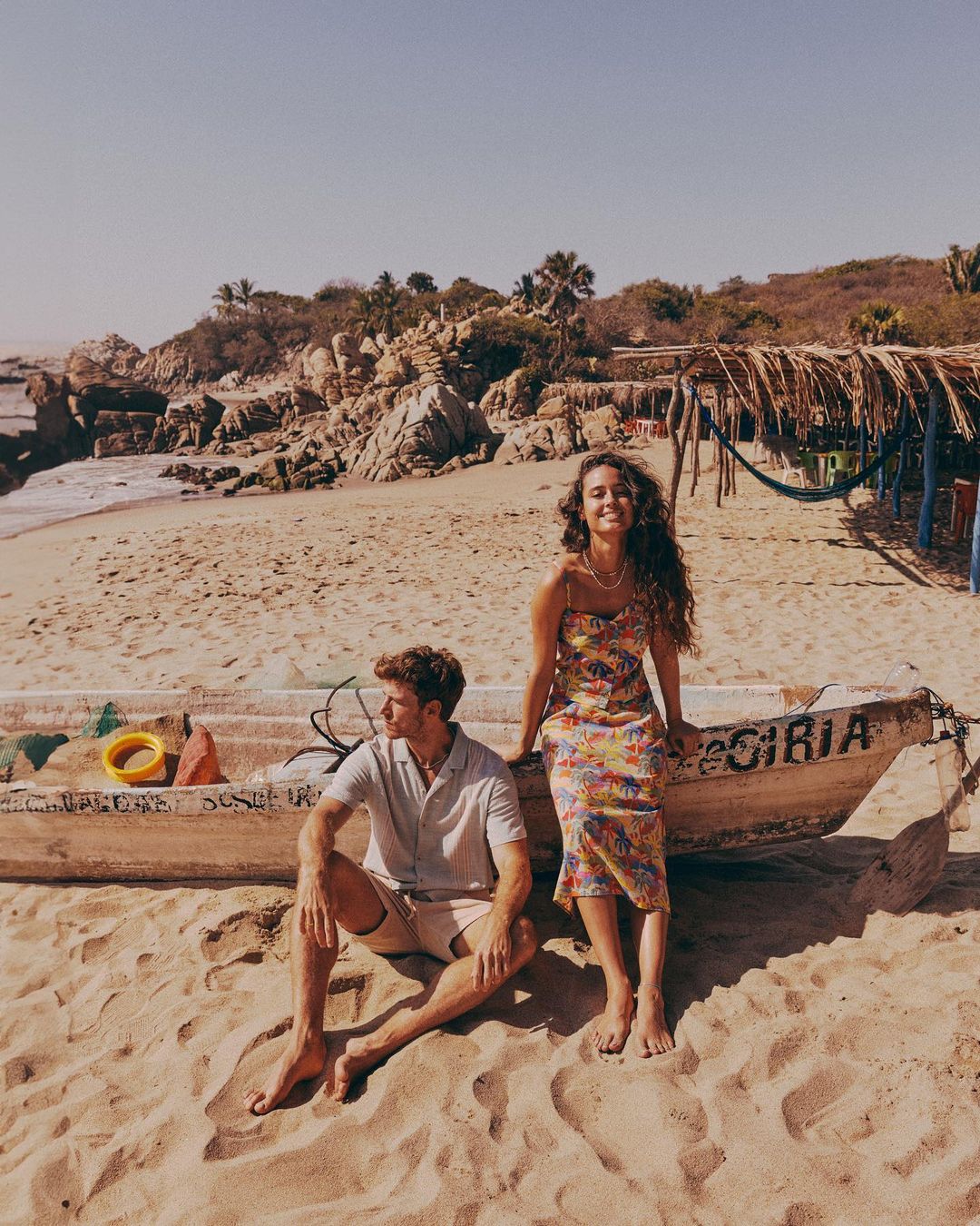 A couple sits on a weathered boat on a sandy beach with tropical foliage. The woman wears a floral dress, and the man is in casual attire.