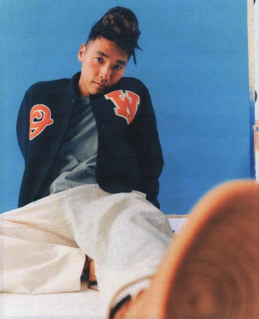 Young Asian man in letterman jacket poses indoors with casual confidence against a blue backdrop, his gaze meeting the viewer's.