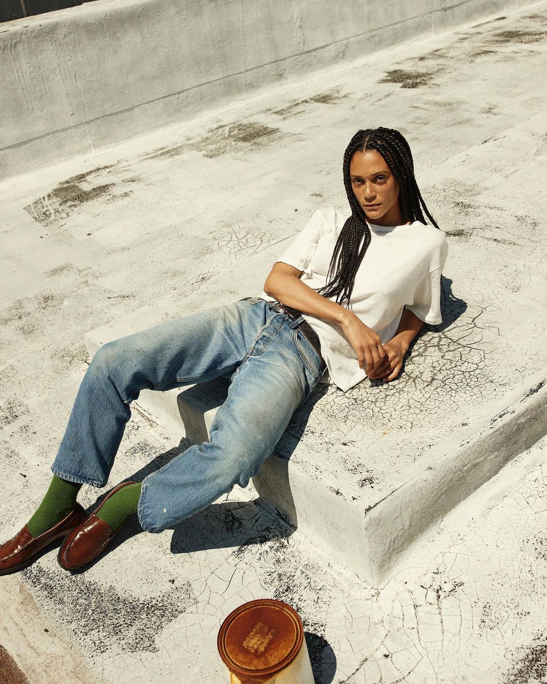Adult woman with braids lounging on a rooftop in a t-shirt and jeans, brown loafers, and green socks poses for a fashion shot.