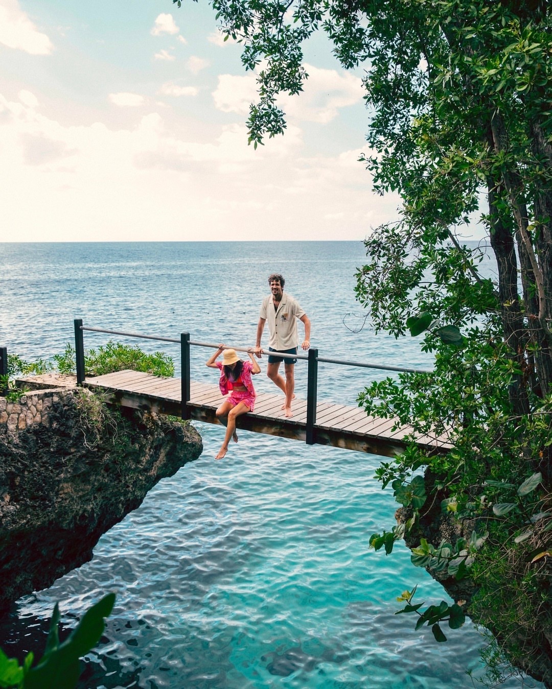 A man and a woman on a small bridge by the ocean. The woman is sitting on the bridge with her legs dangling over the water.