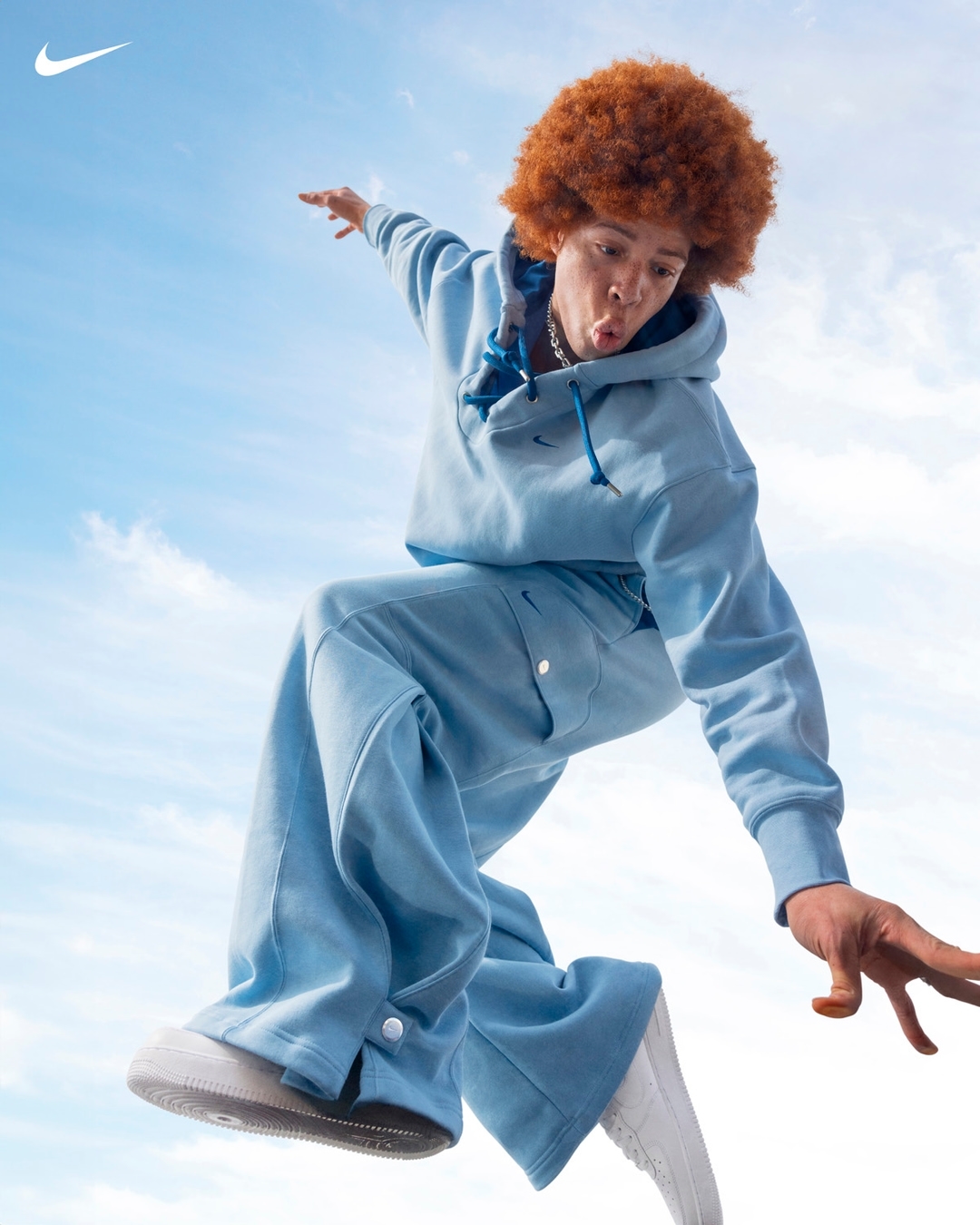 Athletic person in mid-air wearing light blue Nike sweatsuit and white sneakers against a blue sky with clouds, promoting active lifestyle.