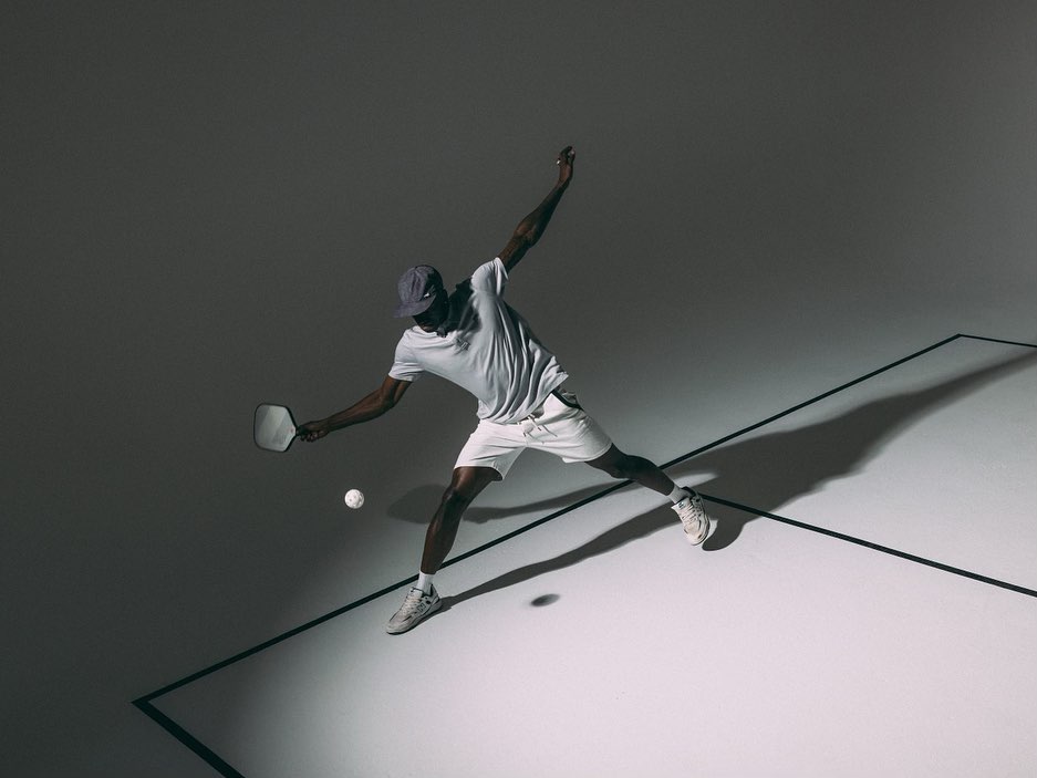 An African-American man is playing pickleball, captured from an eye-level angle in a dynamic pose with a paddle and ball on a grey surface.