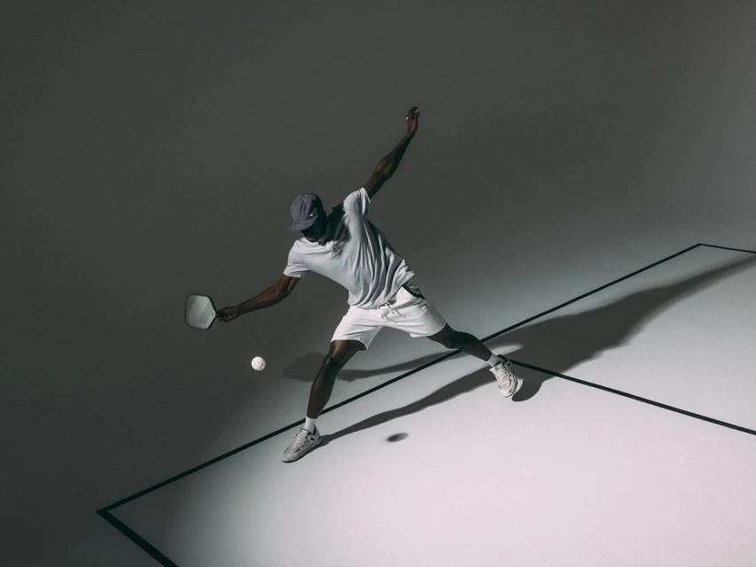 An African-American man is playing pickleball, captured from an eye-level angle in a dynamic pose with a paddle and ball on a grey surface.