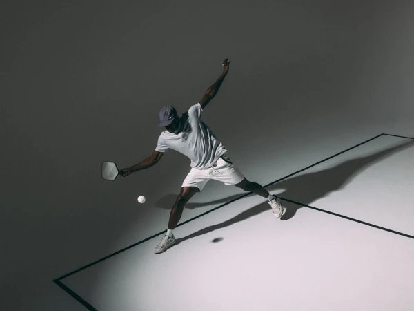 An African-American man is playing pickleball, captured from an eye-level angle in a dynamic pose with a paddle and ball on a grey surface.