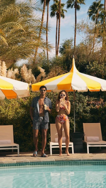 A man and woman in swimwear stand near a swimming pool surrounded by palm trees and umbrellas on a sunny day at a resort.