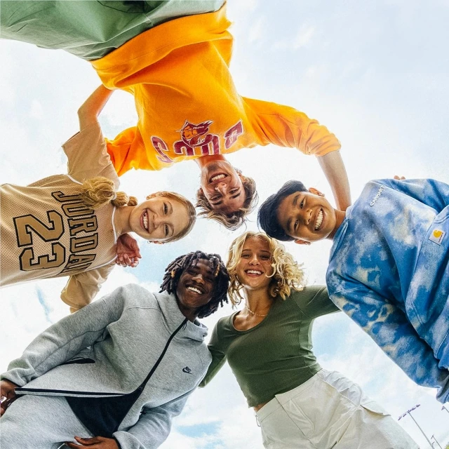 A group of five happy young adults form a circle, smiling and looking down at the camera against a light blue sky backdrop.