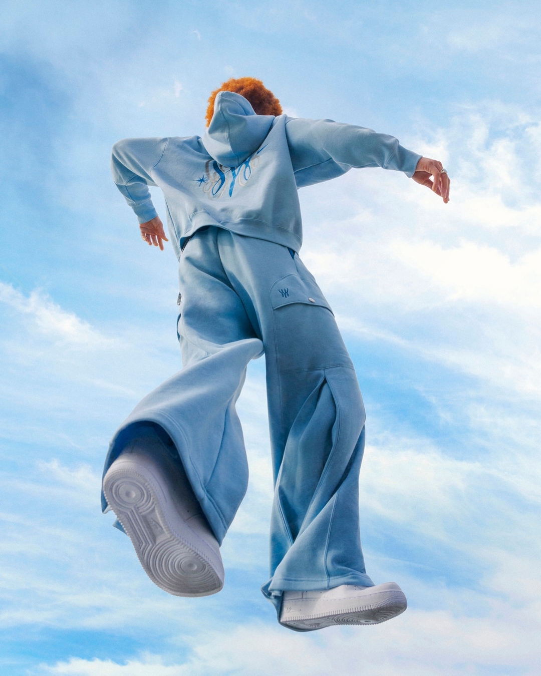 Person in light blue sweatsuit and white sneakers leaps against a blue sky with wispy clouds, seen from a low angle, looking upward.