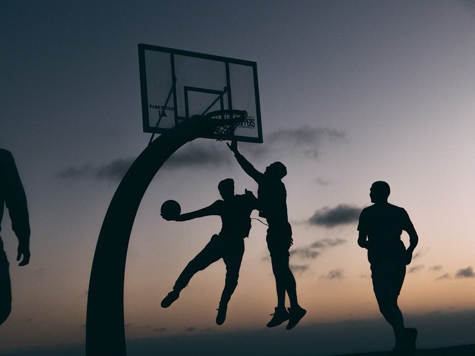 Silhouette of four people playing basketball against a dusky sky with a basketball hoop in action and motion blur.