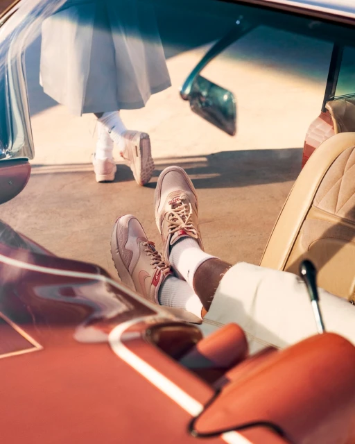 Fashionable scene with tan sneakers inside a vintage car. Woman stands outside with white pleated skirt, mirroring the car's retro vibe.
