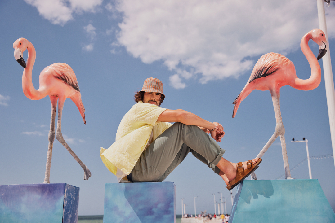 A man sits between two pink flamingo statues on a beach, wearing a yellow shirt, bucket hat, and brown sandals, under a partly cloudy sky.
