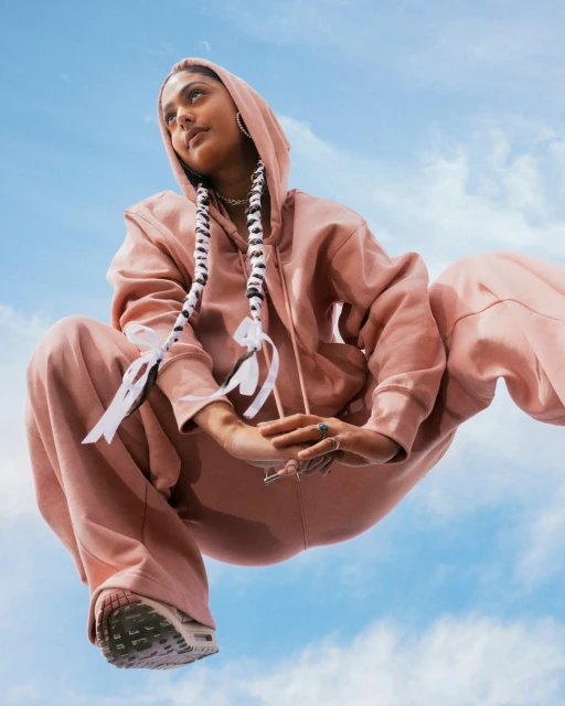 A young adult woman in pink hooded sweatshirt and pants is crouched against a blue sky. Long braids with white ribbons are visible.