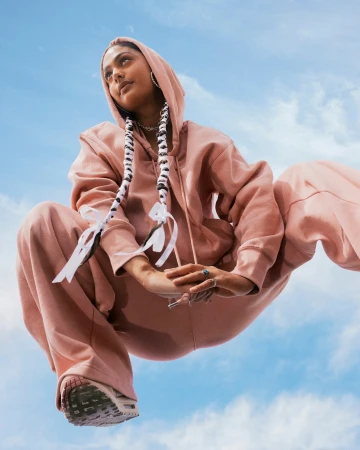 A young adult woman in pink hooded sweatshirt and pants is crouched against a blue sky. Long braids with white ribbons are visible.