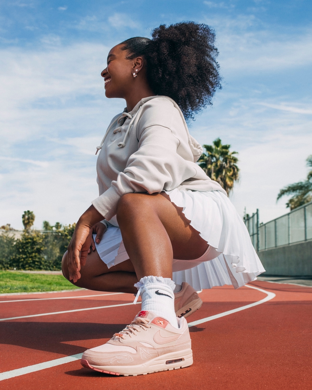 Smiling woman in athletic wear poses on a red running track outdoors under a blue sky, showcasing peach Nike sneakers.