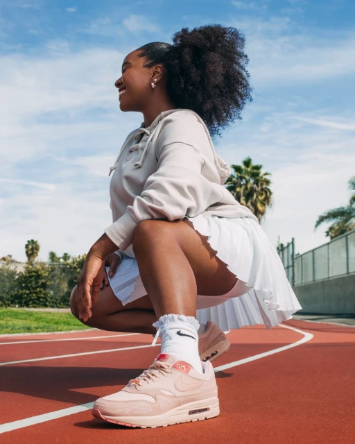 Smiling woman in athletic wear poses on a red running track outdoors under a blue sky, showcasing peach Nike sneakers.