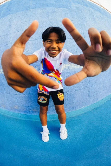 Smiling youth poses in front of a blue wall, framing his face with his hands, wearing shorts and a t-shirt.