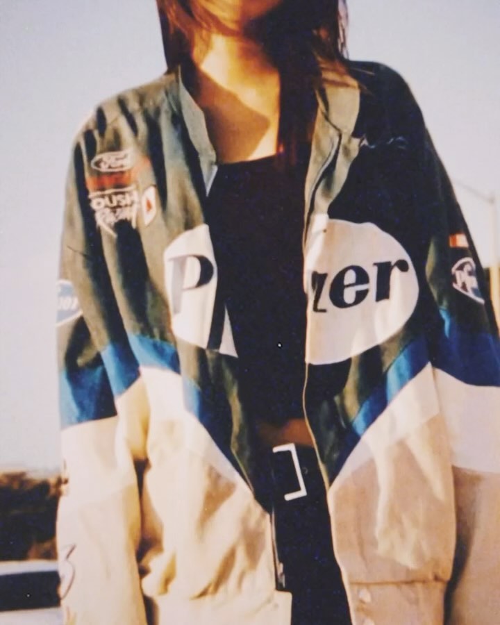 Partial view of a model wearing a vintage racing jacket with visible logos, a black top, and a white belt against an outdoor backdrop.