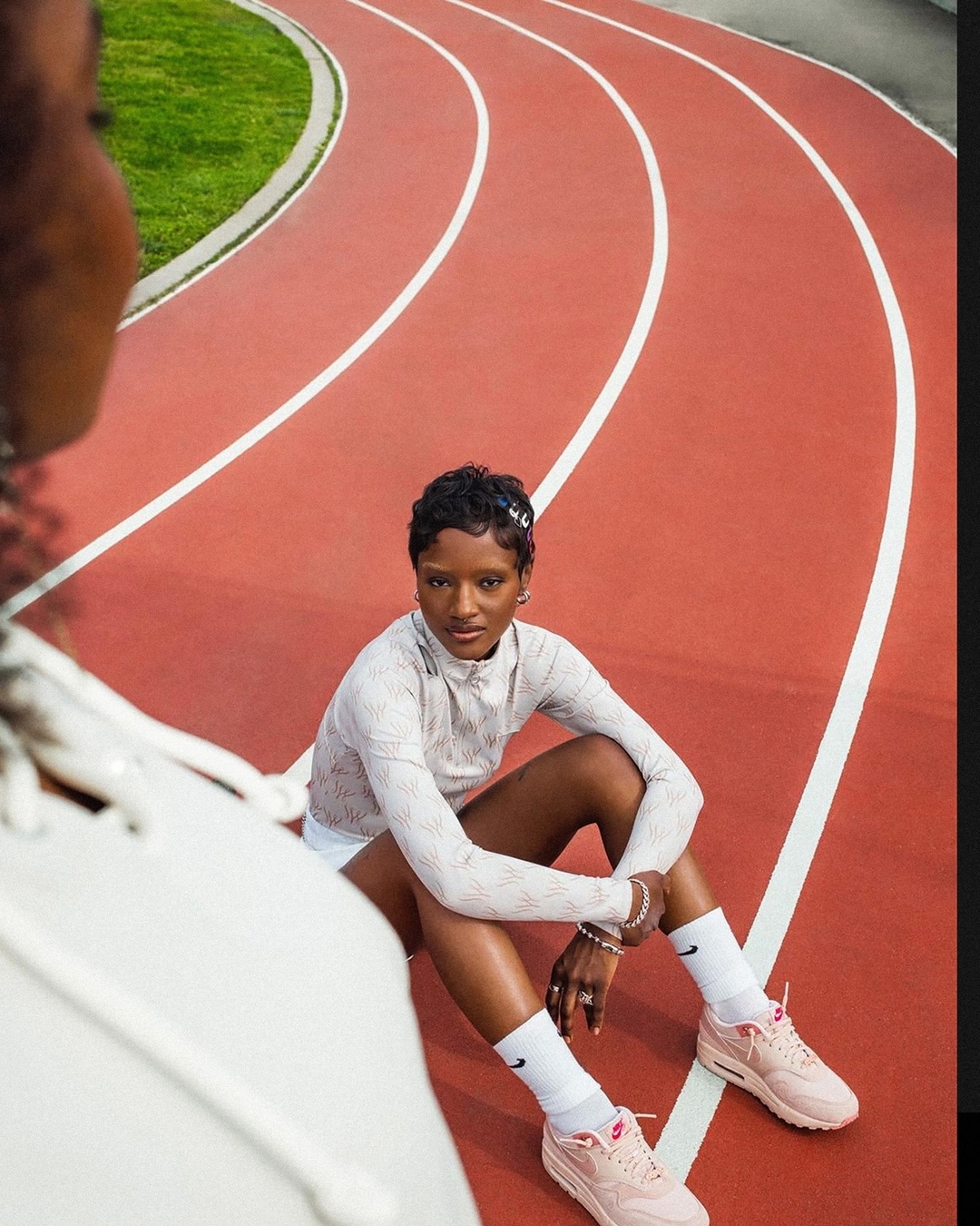 Woman in athletic wear sitting on red running track with green grass in the background, smiling. Outdoor fitness and athletics concept.