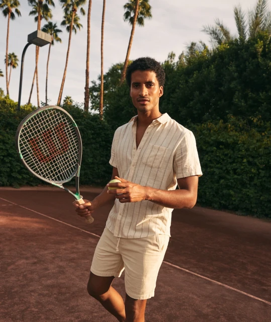 Man in shorts and casual shirt on tennis court holds ball and racquet under palm trees in outdoor setting with warm lighting.