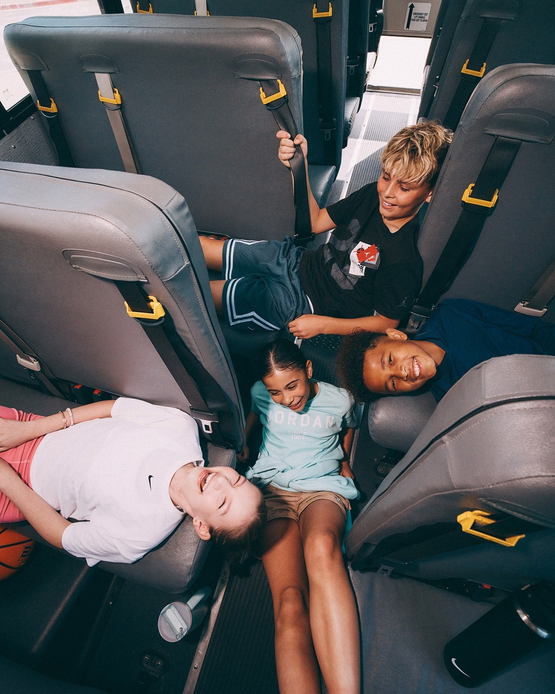 Four children of mixed ethnicity are inside a bus, laying in the aisle laughing, wearing casual clothing, and interacting with each other.
