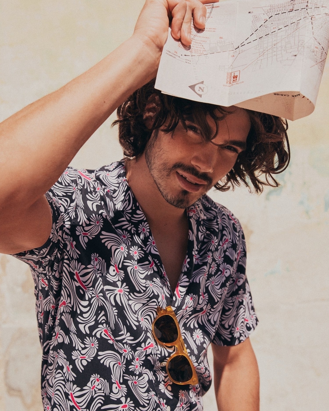 Portrait of a young adult man with curly hair wearing a black and white floral shirt, sunglasses, and holding a map overhead.