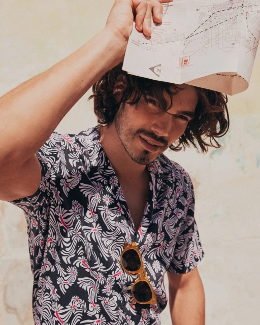 Portrait of a young adult man with curly hair wearing a black and white floral shirt, sunglasses, and holding a map overhead.