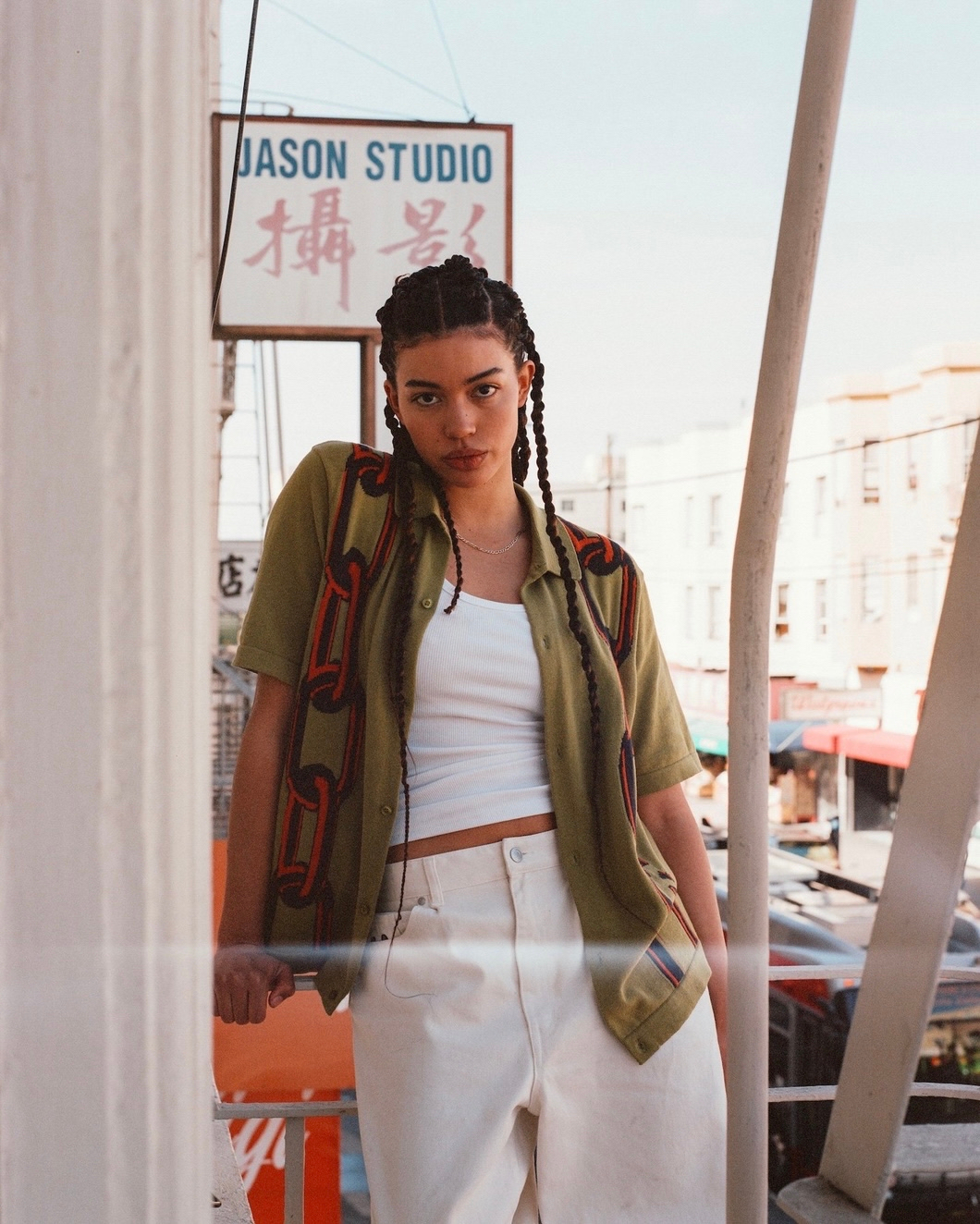 Adult female in a collared shirt stands outside Jason Studio. She wears white jeans and poses in front of buildings in daylight.