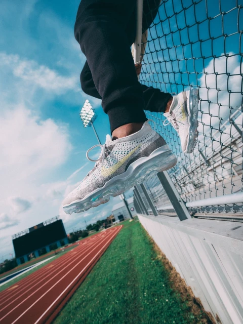 Low angle of person sitting on fence near track wearing athletic shoes with blue sky and clouds in the background on a sunny day.