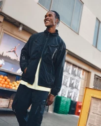 Black man smiling outdoors in New York wearing a dark jacket over a pale yellow t-shirt and dark cargo pants by Arturo Torres