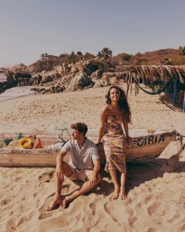 Man sitting in sand and woman sitting on weathered wooden boat on a sunny beach