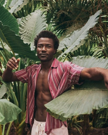 Black man wearing an open striped short-sleeve shirt among lush tropical green leaves