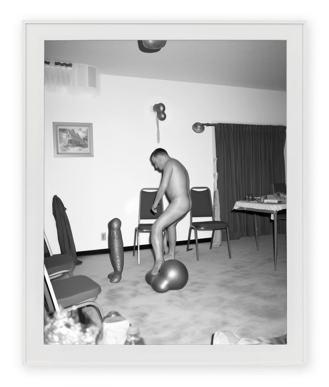 Black and white photograph of an adult male standing on a large rubber ball next to an inflatable figure in an indoor setting.