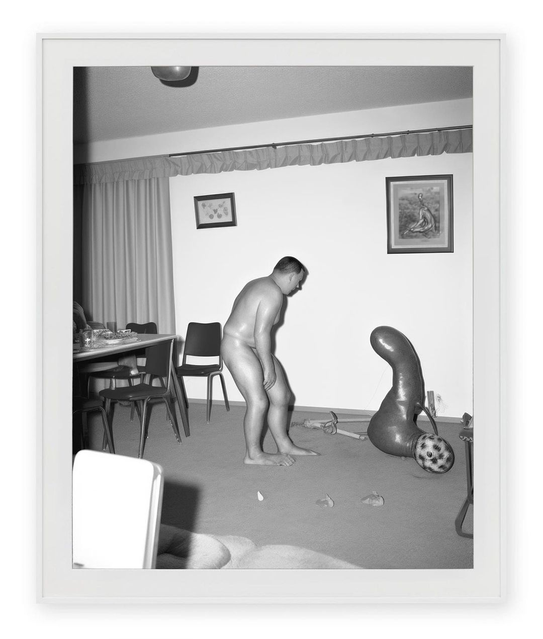 Monochrome image of a shirtless adult man standing in an indoor setting next to a balloon and table with chairs.