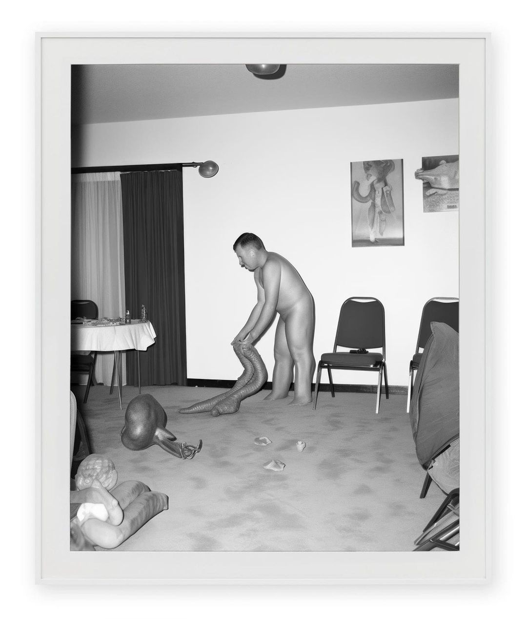 Monochrome photograph of a nude adult male interacting with a snake-like creature, set in an indoor room with chairs and artwork.