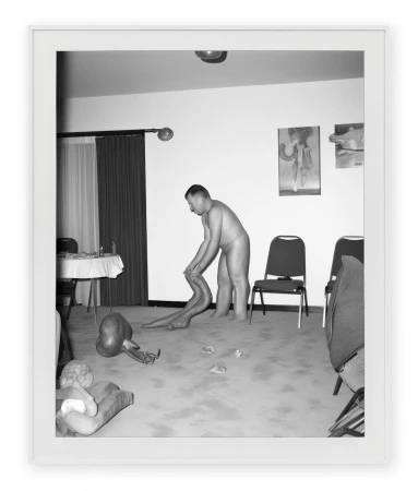 Monochrome photograph of a nude adult male interacting with a snake-like creature, set in an indoor room with chairs and artwork.