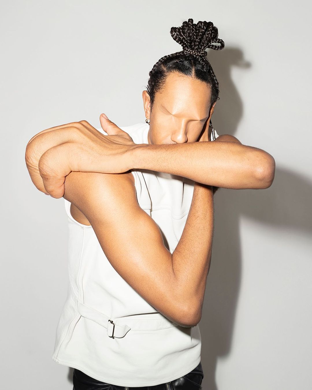 Man in a white vest poses with arms crossed, face obscured, and hair in a bun, set against a neutral gray studio backdrop.