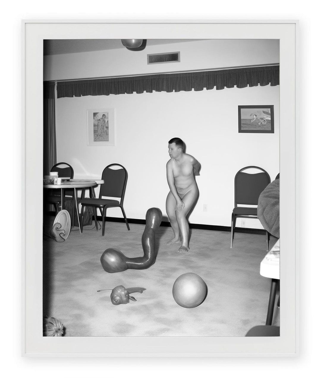 Black and white photograph in a white frame showing an adult posing near abstract sculptures and furniture inside an indoor room.