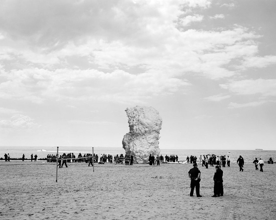 Black and white shot of large group of people gathered on a sandy beach by a rock-like sculpture under a cloudy sky near the ocean.