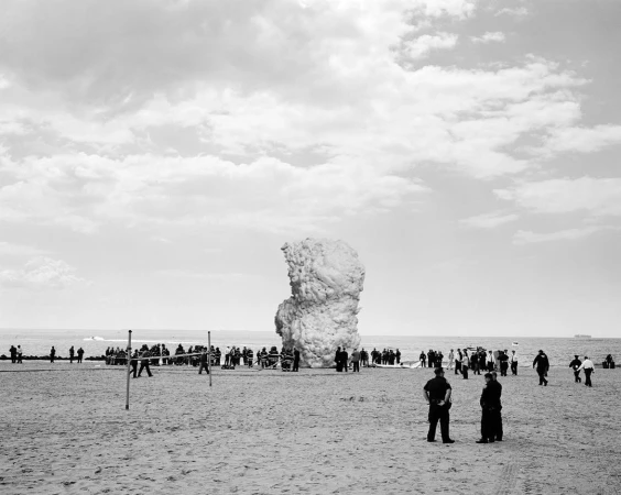 Black and white shot of large group of people gathered on a sandy beach by a rock-like sculpture under a cloudy sky near the ocean.