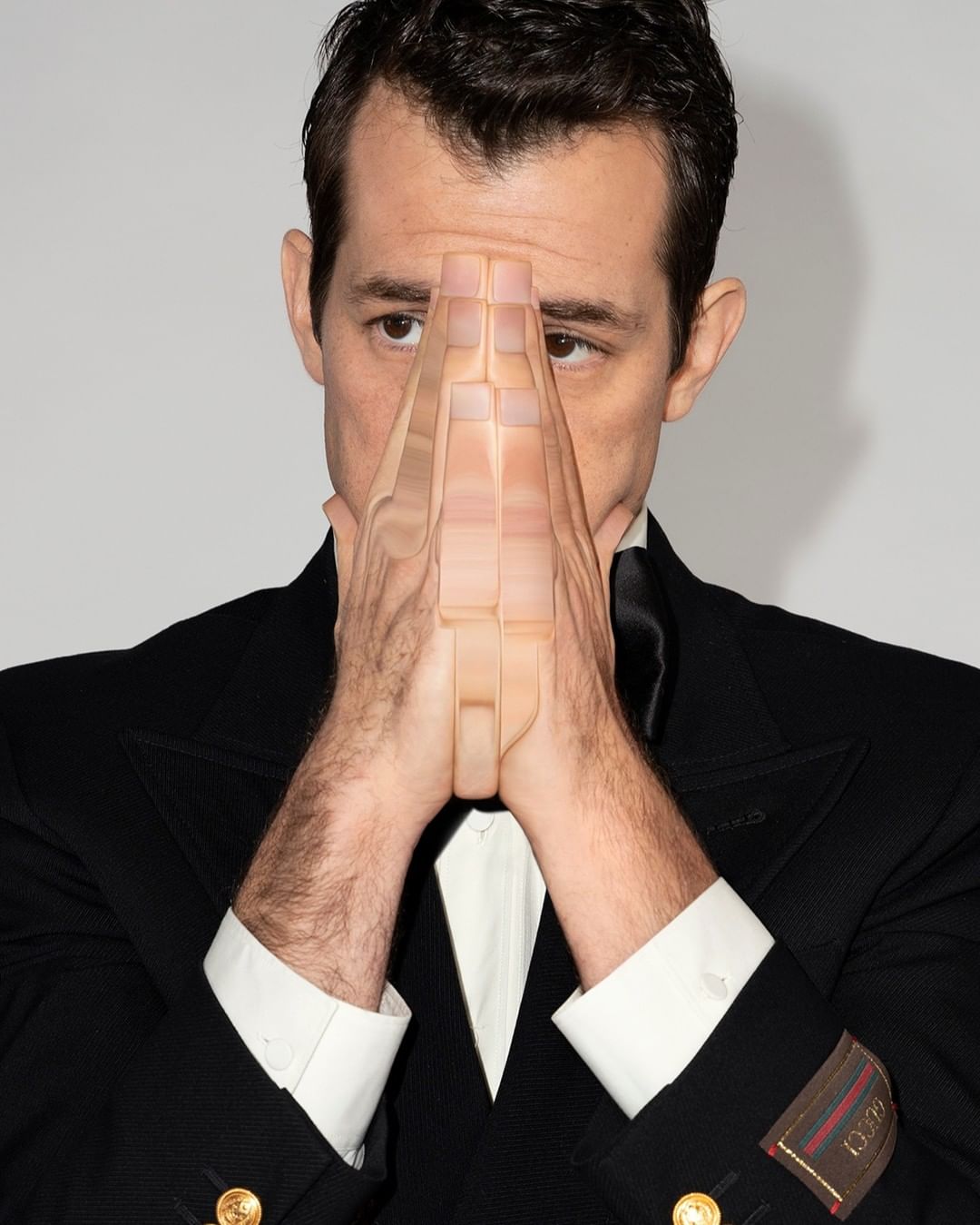 Headshot of an adult male in a black suit with hands near his face, photographed in a photo studio against a neutral grey backdrop.