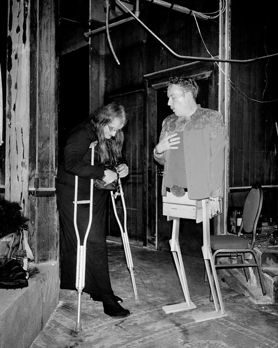 Black and white image of a woman adjusting crutches next to a man with a wooden prosthetic lower body on a stage indoors.