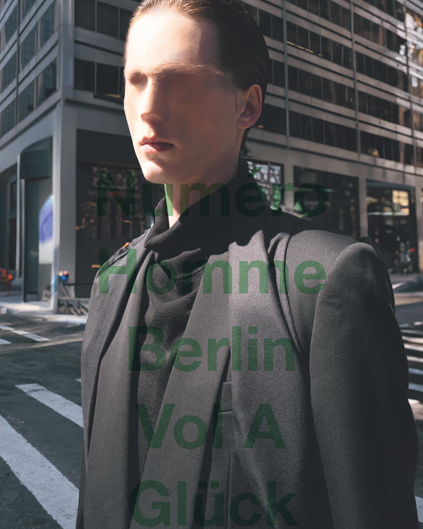 Man in a dark suit stands on a city street in a fashion editorial, featuring modern architecture and natural sunlight.