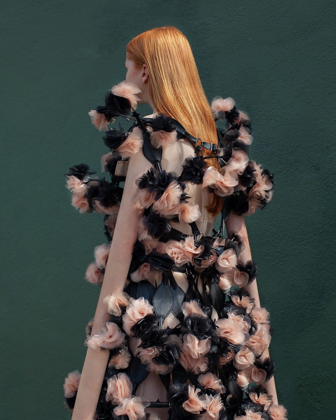 Back view of a woman with red hair wearing a floral dress against a green backdrop, showcasing fashion and editorial style.