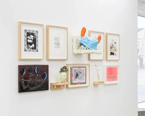 Installation view of framed artworks and sculpture pieces hanging on a white wall inside of a gallery space with natural lighting.