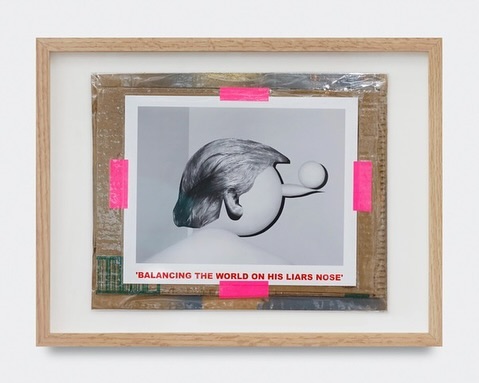 A framed photograph shows a sculptural head with gray hair balancing a white ball on its nose, secured by pink tape on cardboard backing.